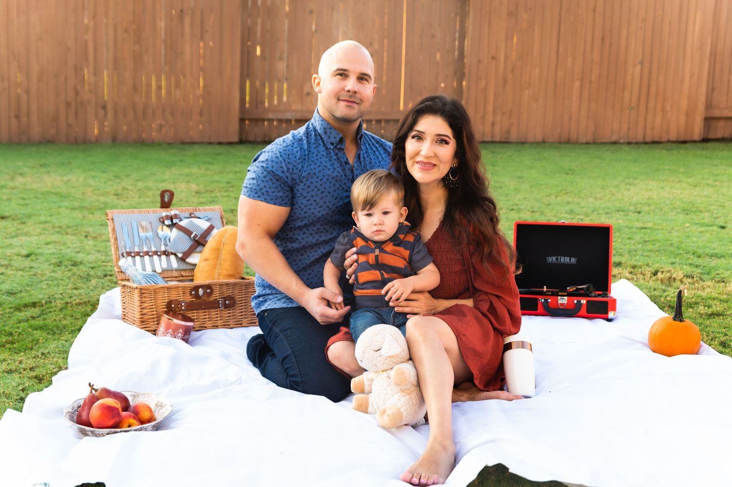 Family having picnic on white blanket in backyard with wooden fence.