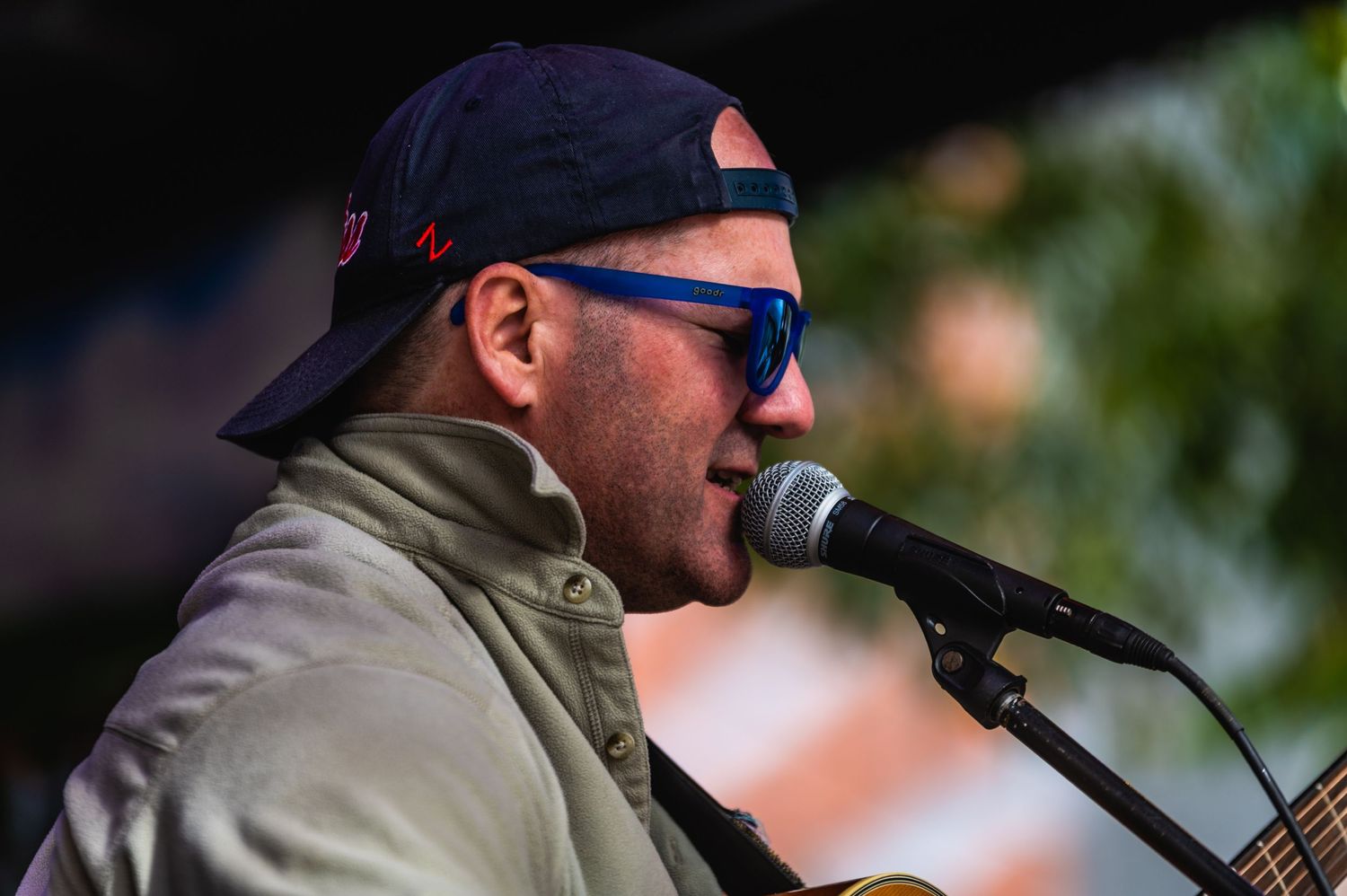 Singer wearing baseball cap and sunglasses performs into microphone during outdoor concert.