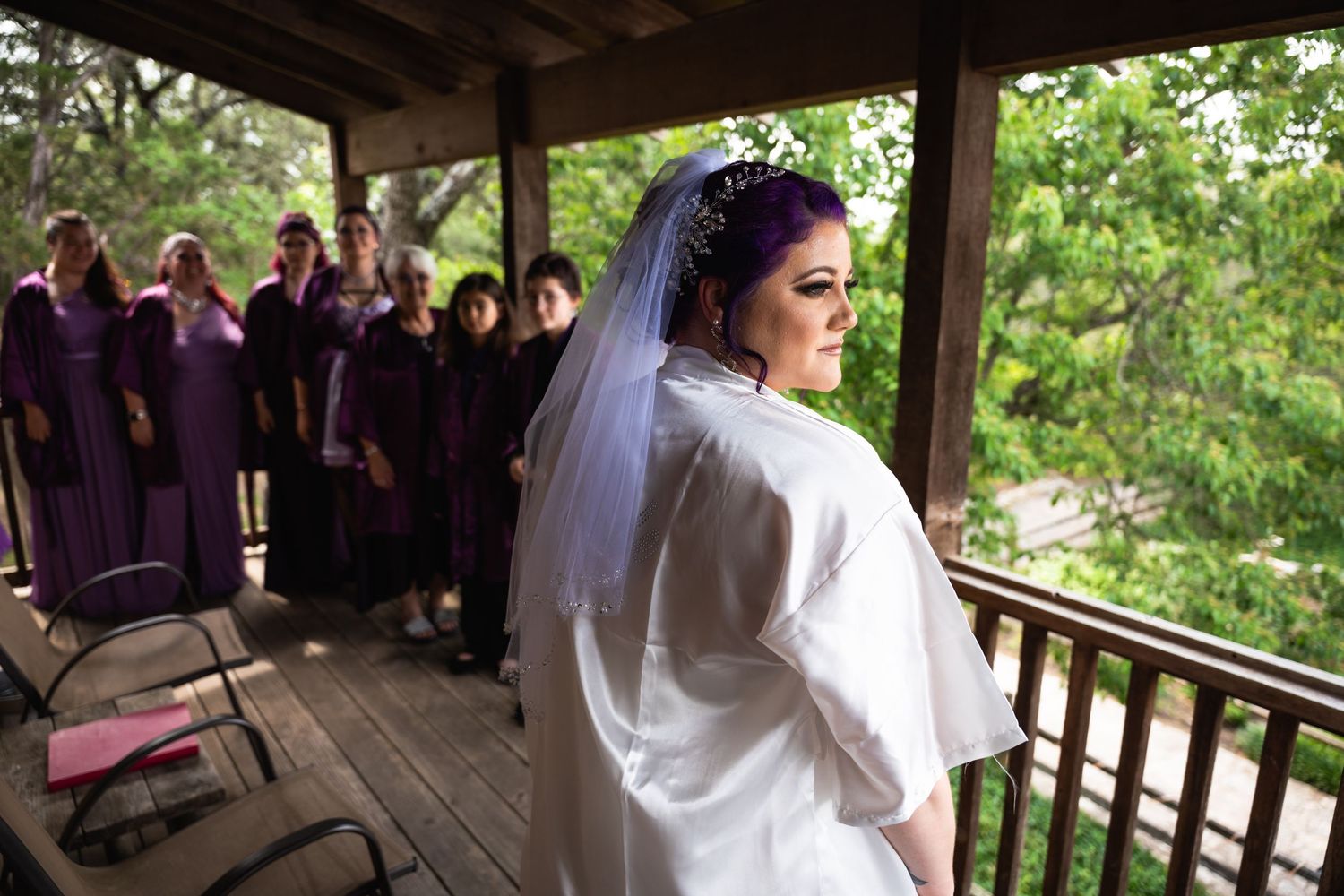 Bride in white robe stands on porch with bridesmaids in purple dresses behind her.
