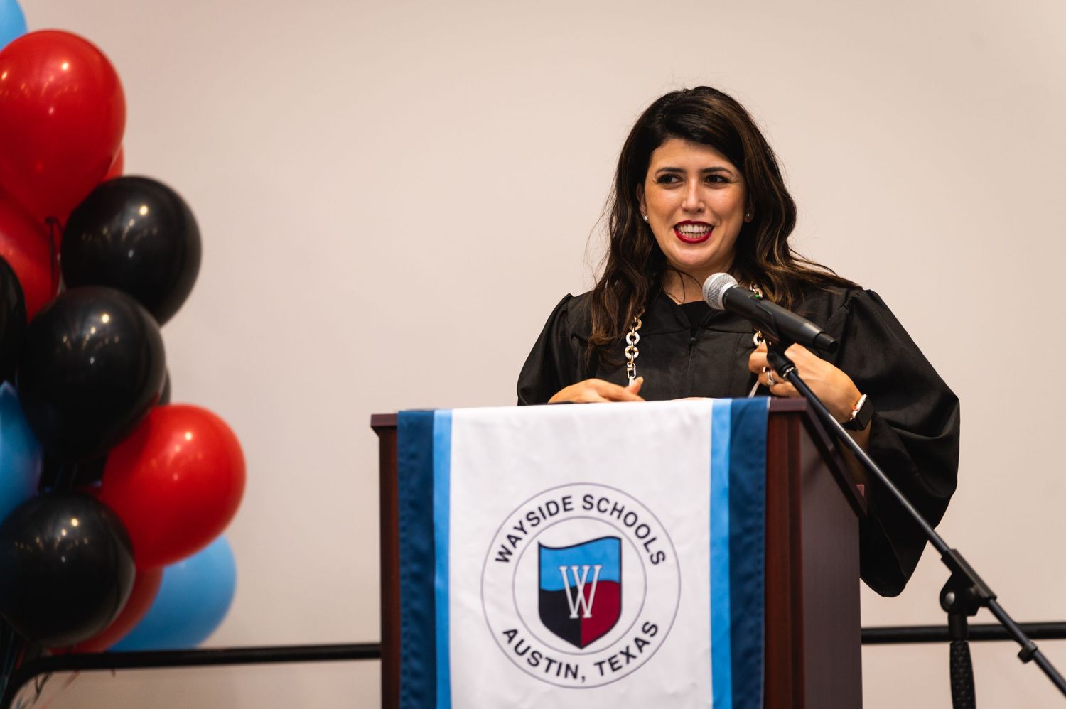 Graduate delivering speech at podium with red and black balloons in background.