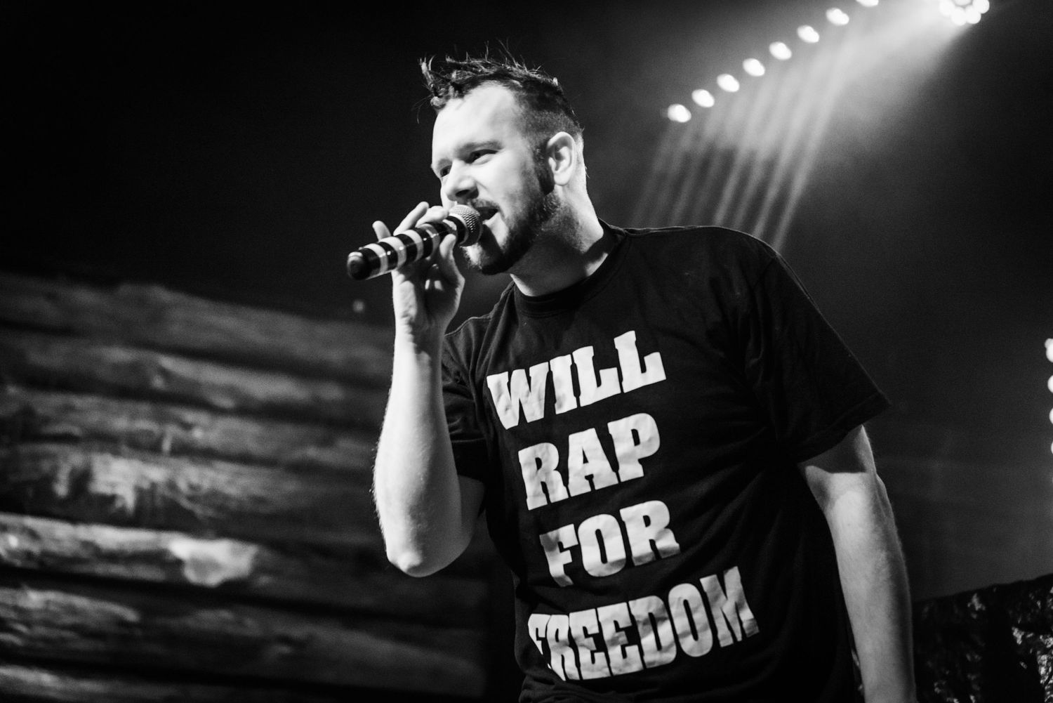 Black and white concert photo of performer in 'Will Rap For Freedom' t-shirt on stage.