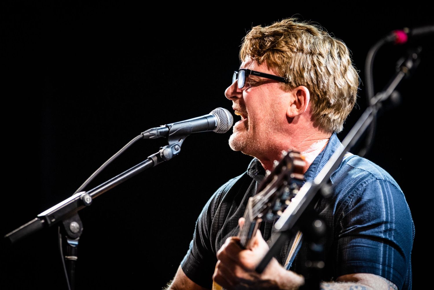 Musician in denim shirt singing into microphone while playing guitar on dark stage.