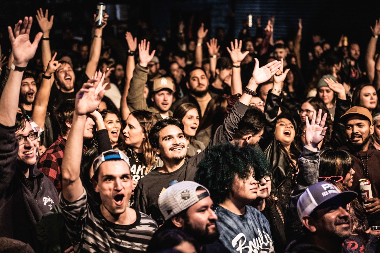An energetic crowd raises their hands at a concert with people packed tightly together.