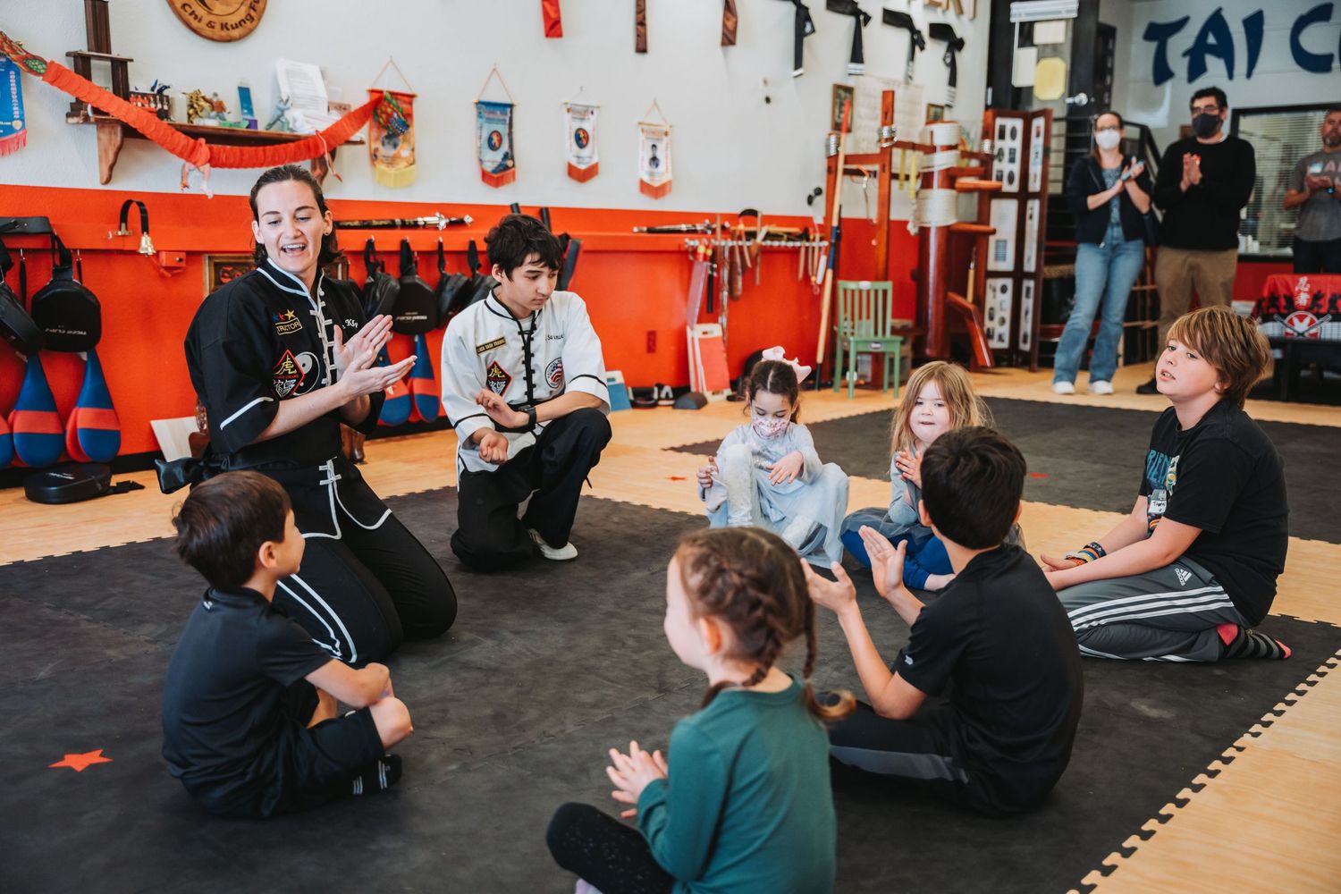 Martial arts training session on black mats with orange walls and equipment in the background.
