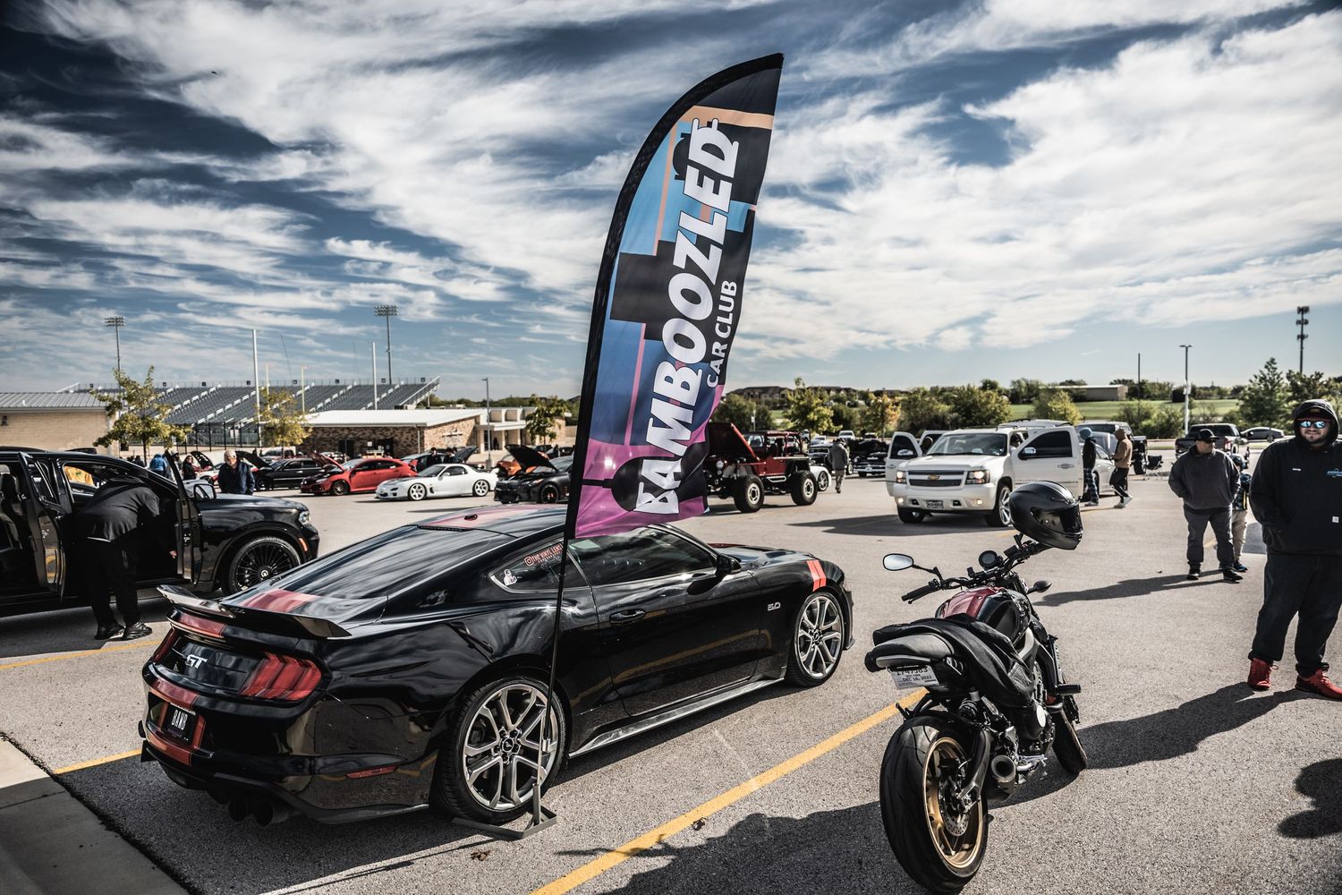 Black Mustang and motorcycle displayed at automotive event under cloudy sky.