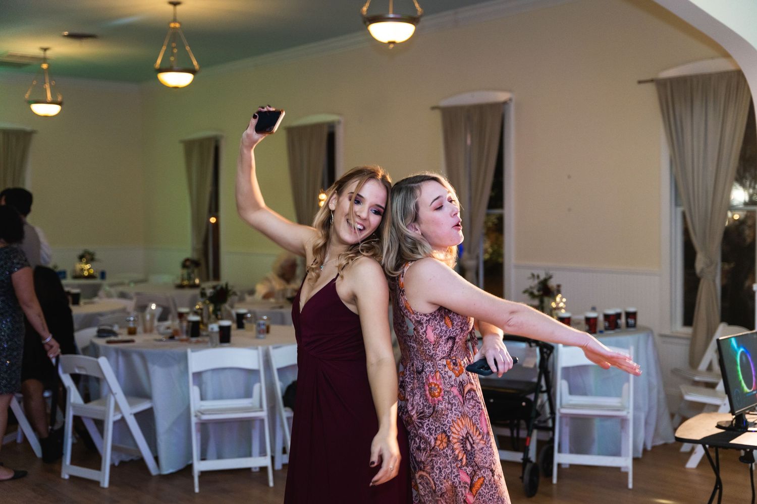 Two wedding guests strike fun poses while dancing in a reception hall with table settings visible.