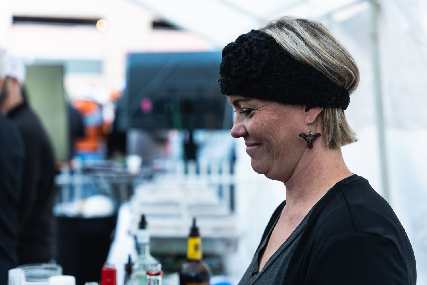 Person in black clothing and headwear smiling while working at an outdoor beverage station.