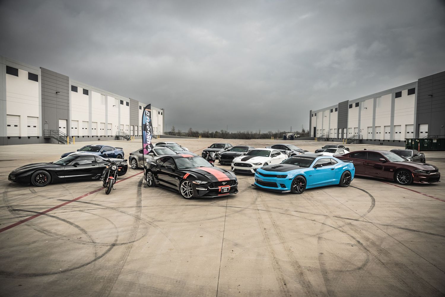 High-performance cars displayed in formation against industrial backdrop with cloudy skies.