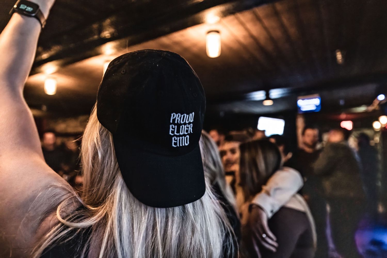 Concert attendee wearing black baseball cap with white text shown from behind in dimly lit venue.