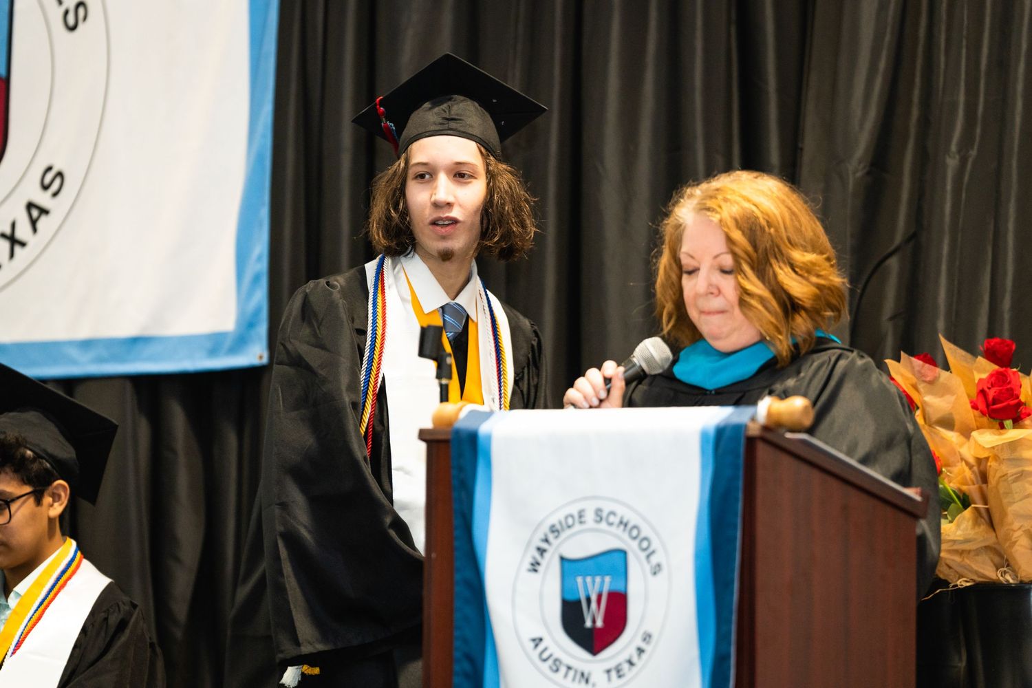Graduate receiving diploma at commencement ceremony with school banner in background.