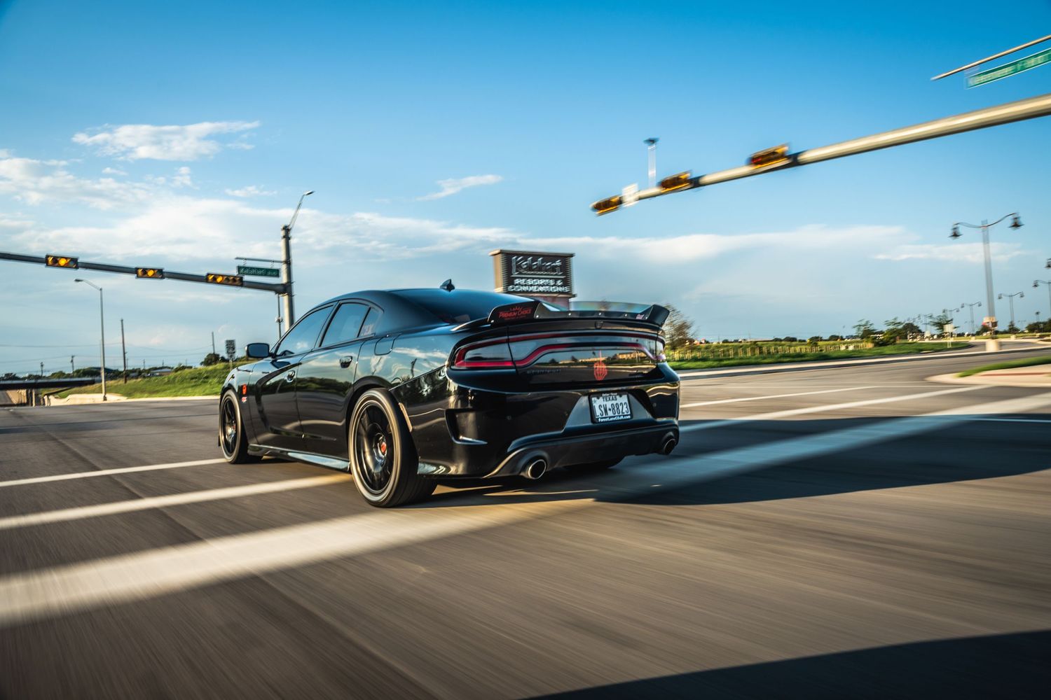 Black Dodge Charger with custom wheels cruising on highway under blue sky.