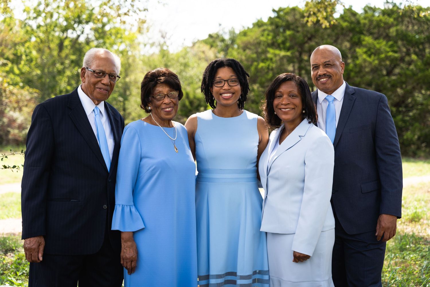 Group of five adults in formal attire pose together against natural backdrop.