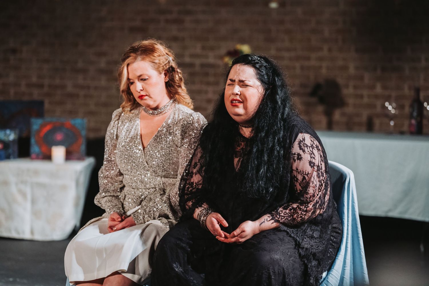 Two performers share a scene while seated on stage in formal attire.