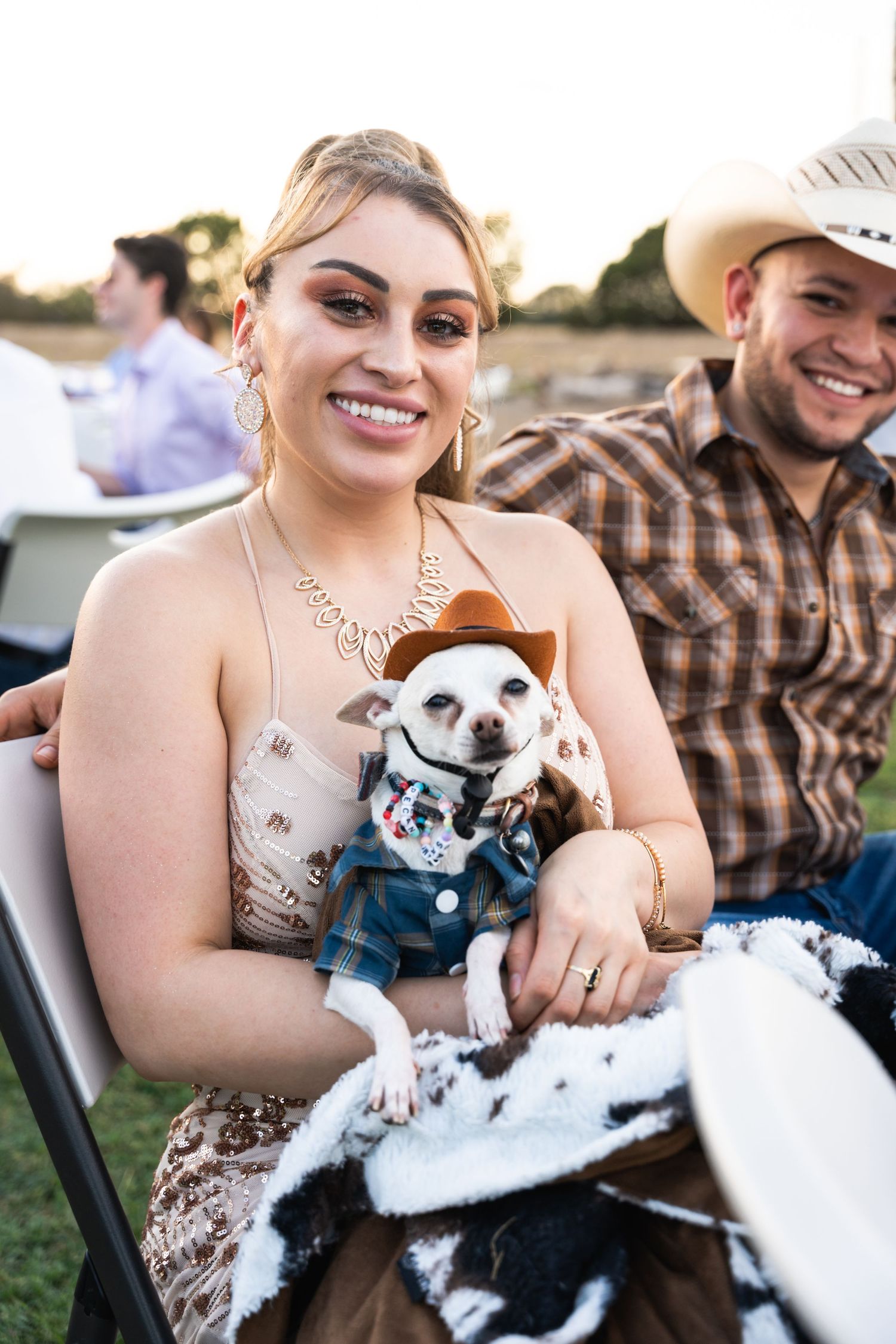 Small dog wearing cowboy outfit and formal wear sits with guests at outdoor wedding reception.