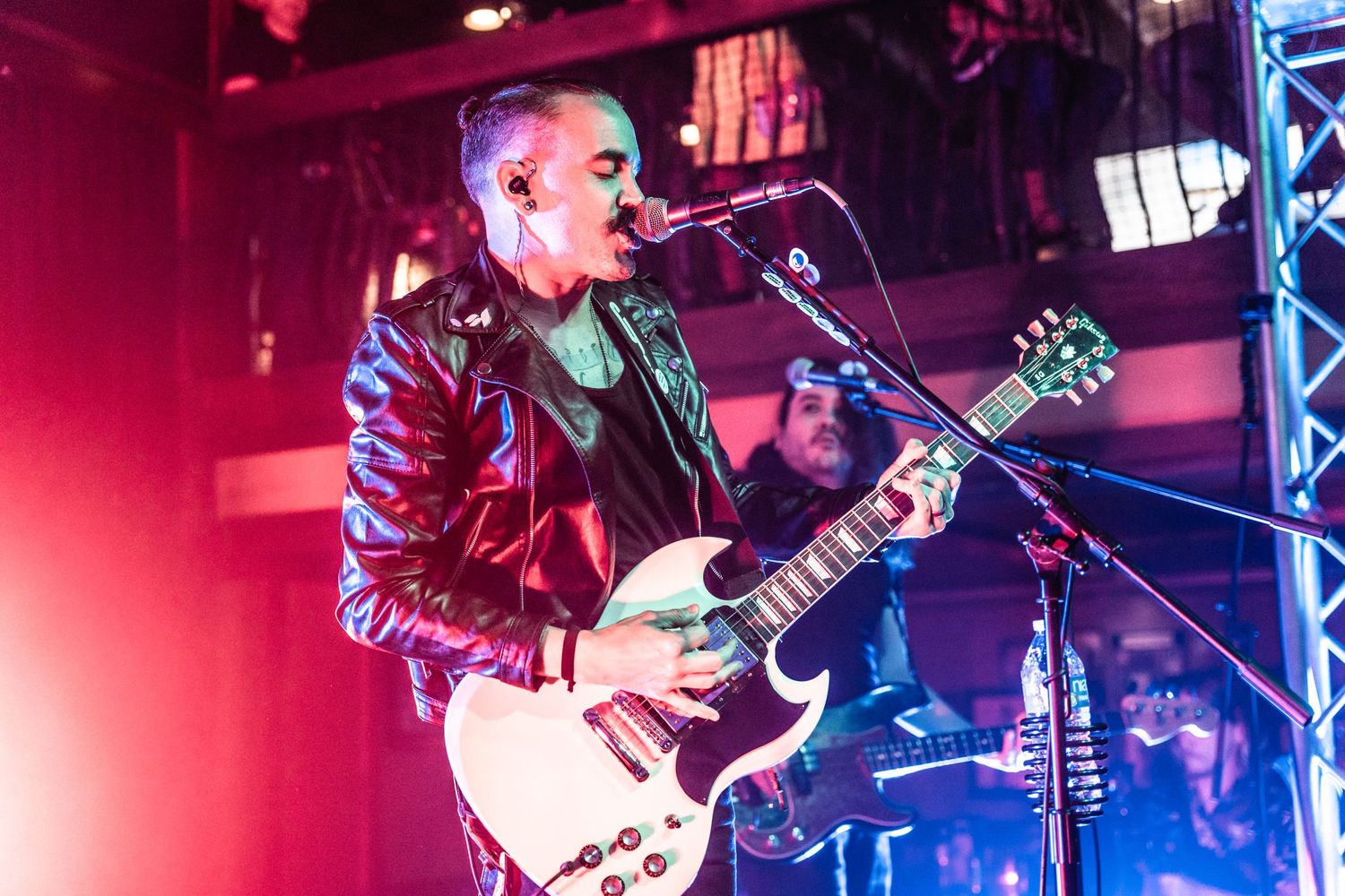Musician in leather jacket performs passionately on guitar under dramatic red stage lighting.