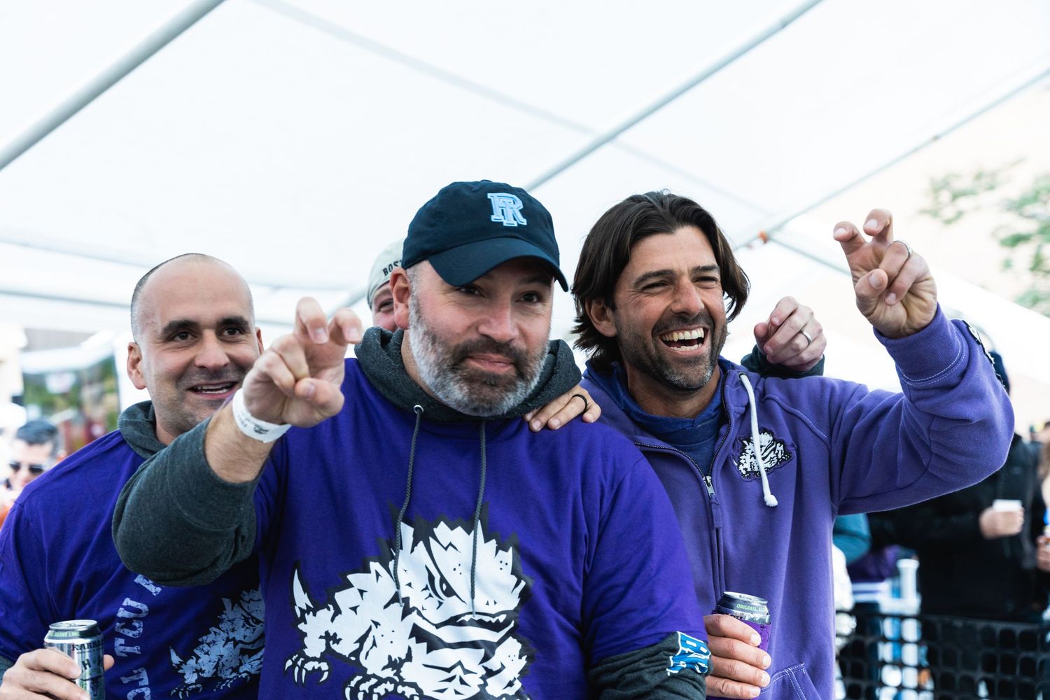 Group of friends in purple apparel cheering and raising drinks at a tailgate party.