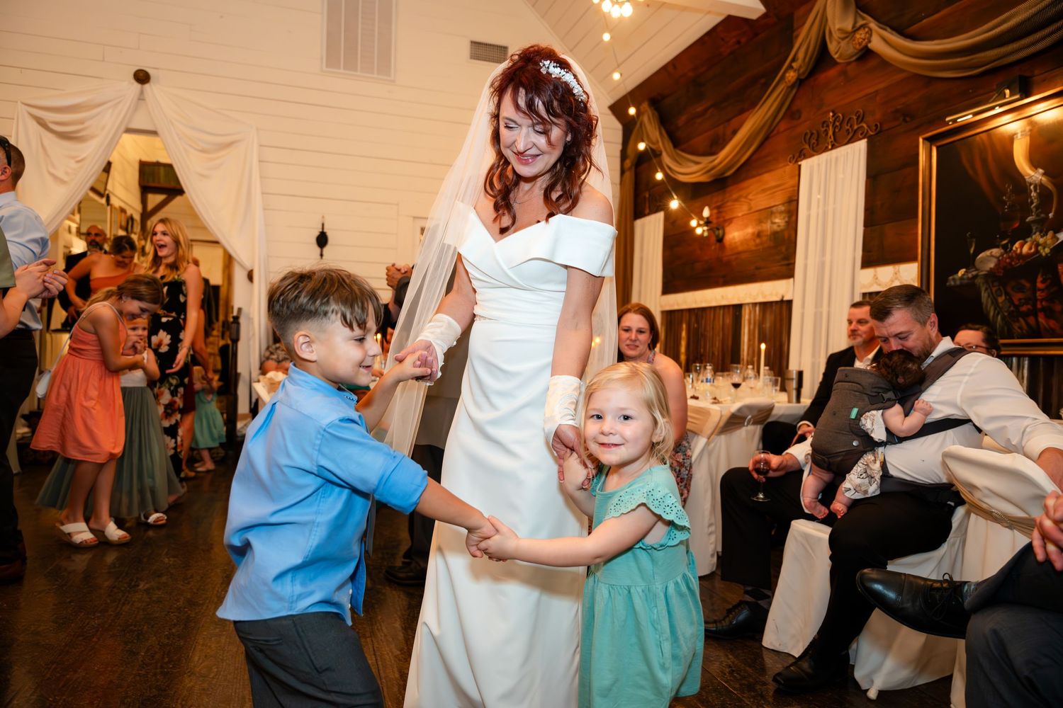 Bride in white dress dancing with young children at a wedding reception.