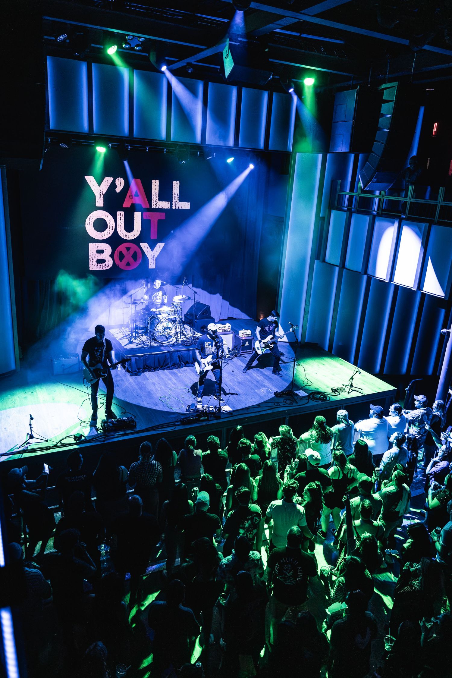 Concert venue filled with fans watching Y'all Out Boy perform under dramatic blue and green lighting.