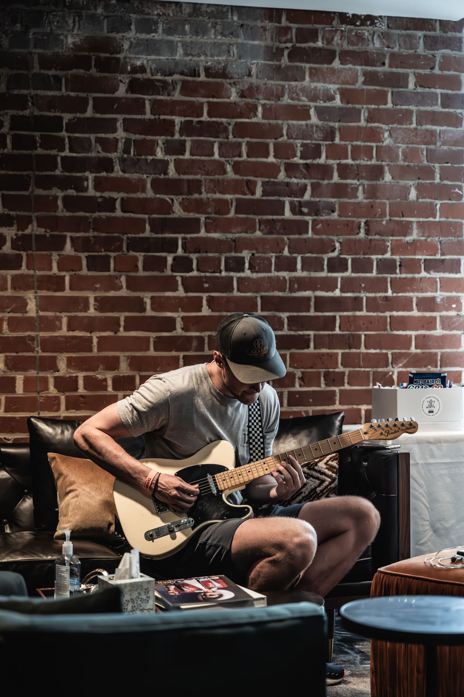 Musician playing electric guitar while seated against exposed brick wall in intimate setting.