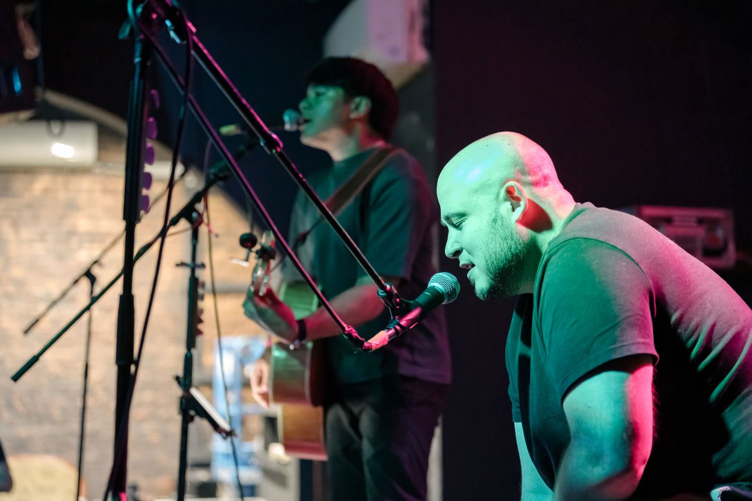 Musicians performing with acoustic guitar and vocals under green stage lighting at intimate venue.