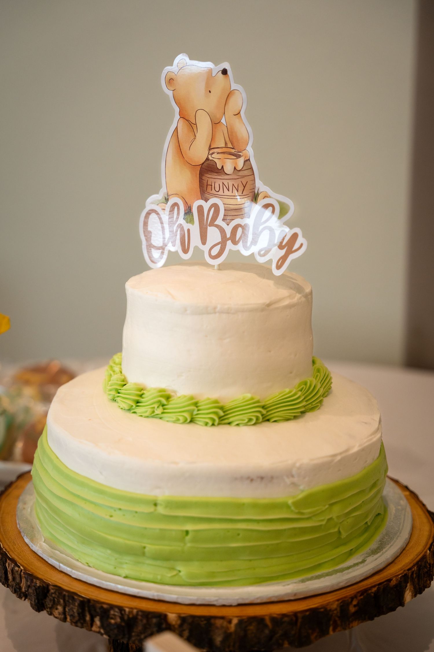 Two-tiered white and green baby shower cake with Winnie the Pooh themed topper on wooden stand.