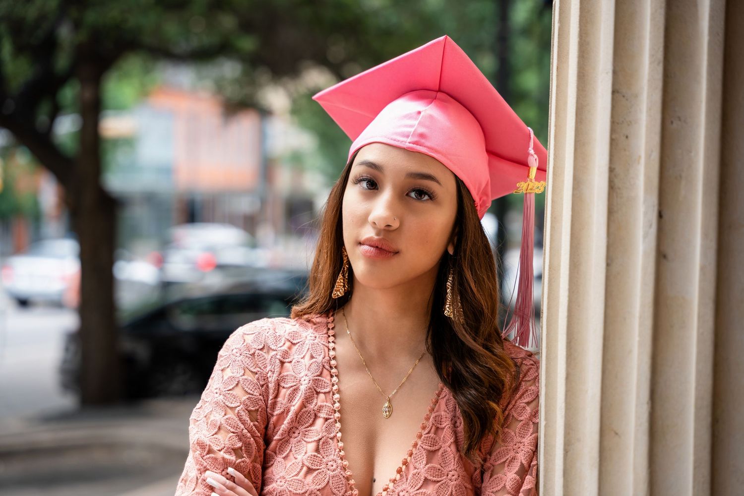 Graduate in pink cap and lace dress poses next to stone column in urban setting.