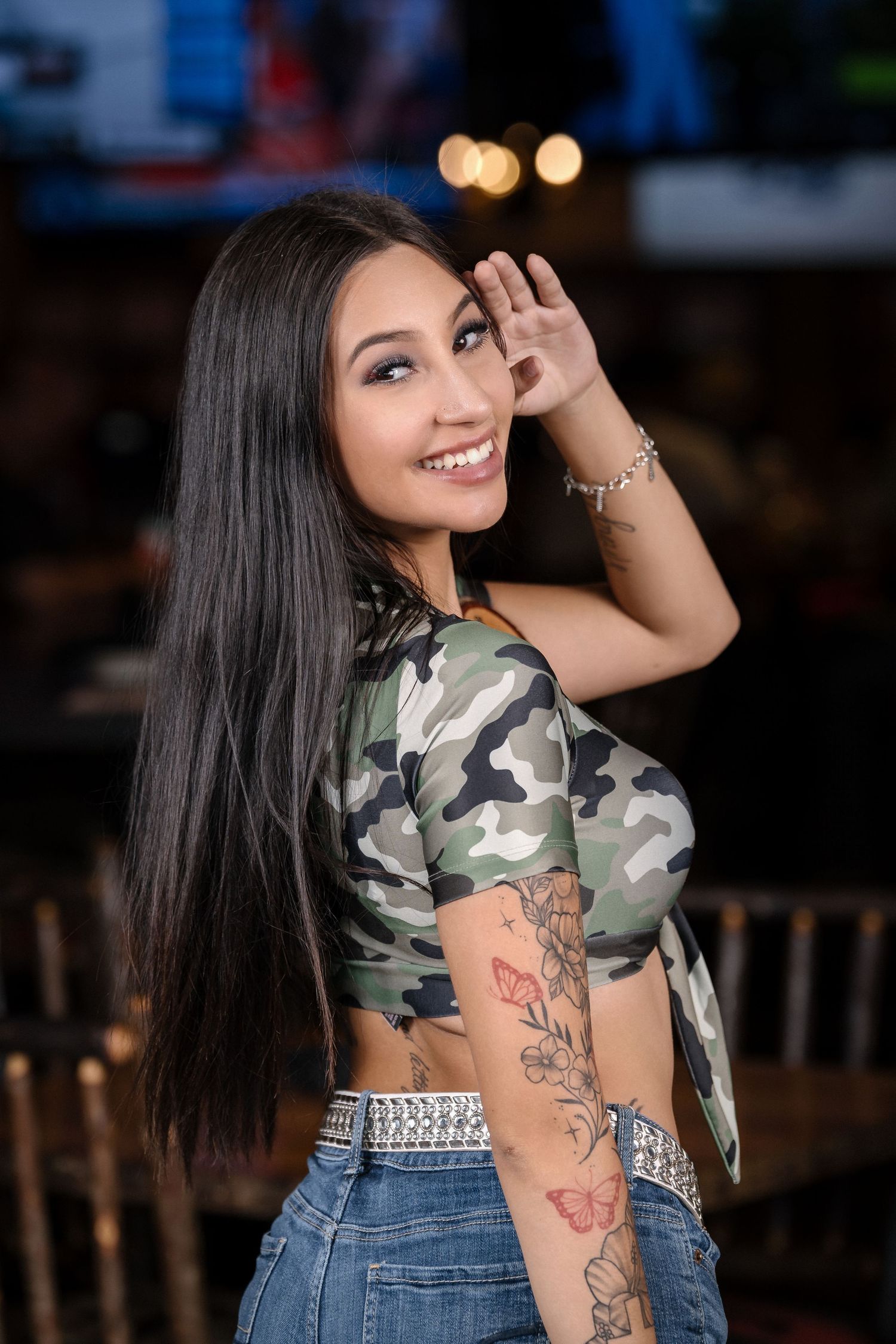 Model wearing camo crop top and jeans poses with long dark hair against blurred background.