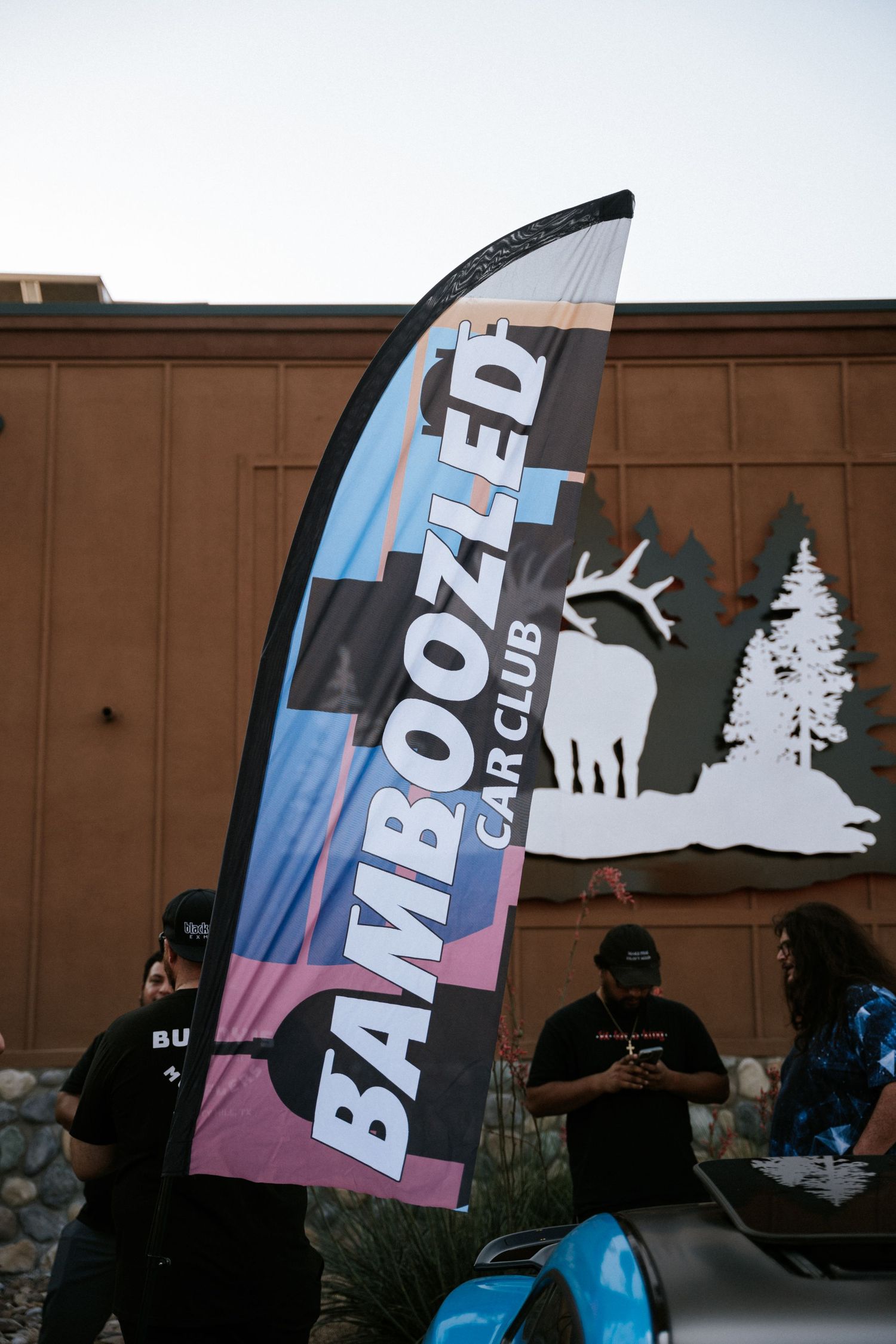 Bamboozled Car Club feather flag banner against brown building exterior.