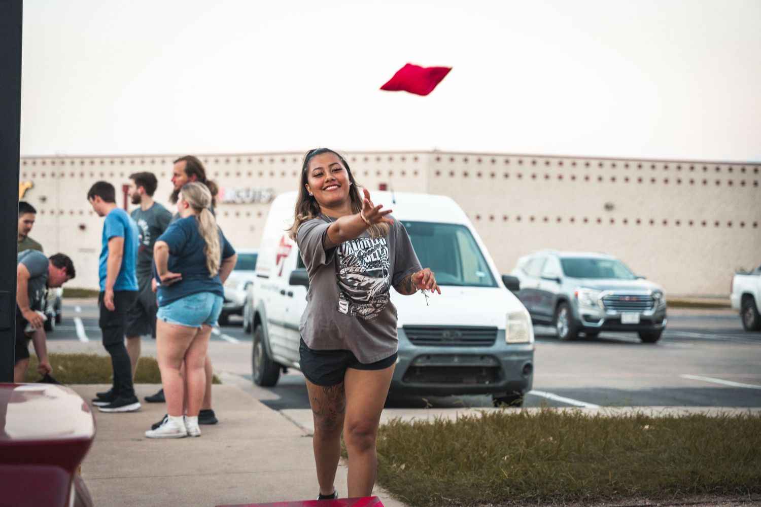 Red cornhole bag being tossed during outdoor game in parking lot.