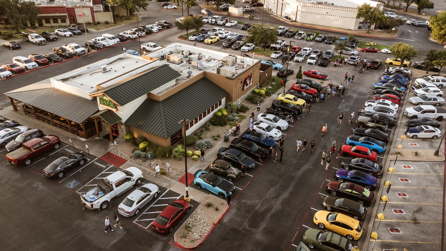Bird's eye view of a shopping center parking lot filled with cars at a car meet.