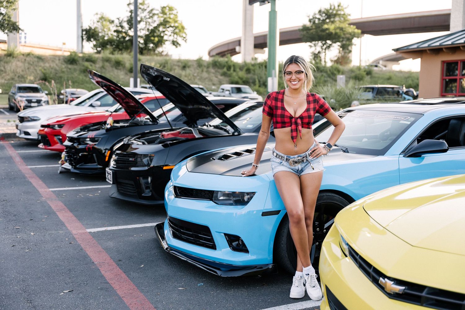 Person in casual attire poses with line of performance cars including Camaros at outdoor car meet.