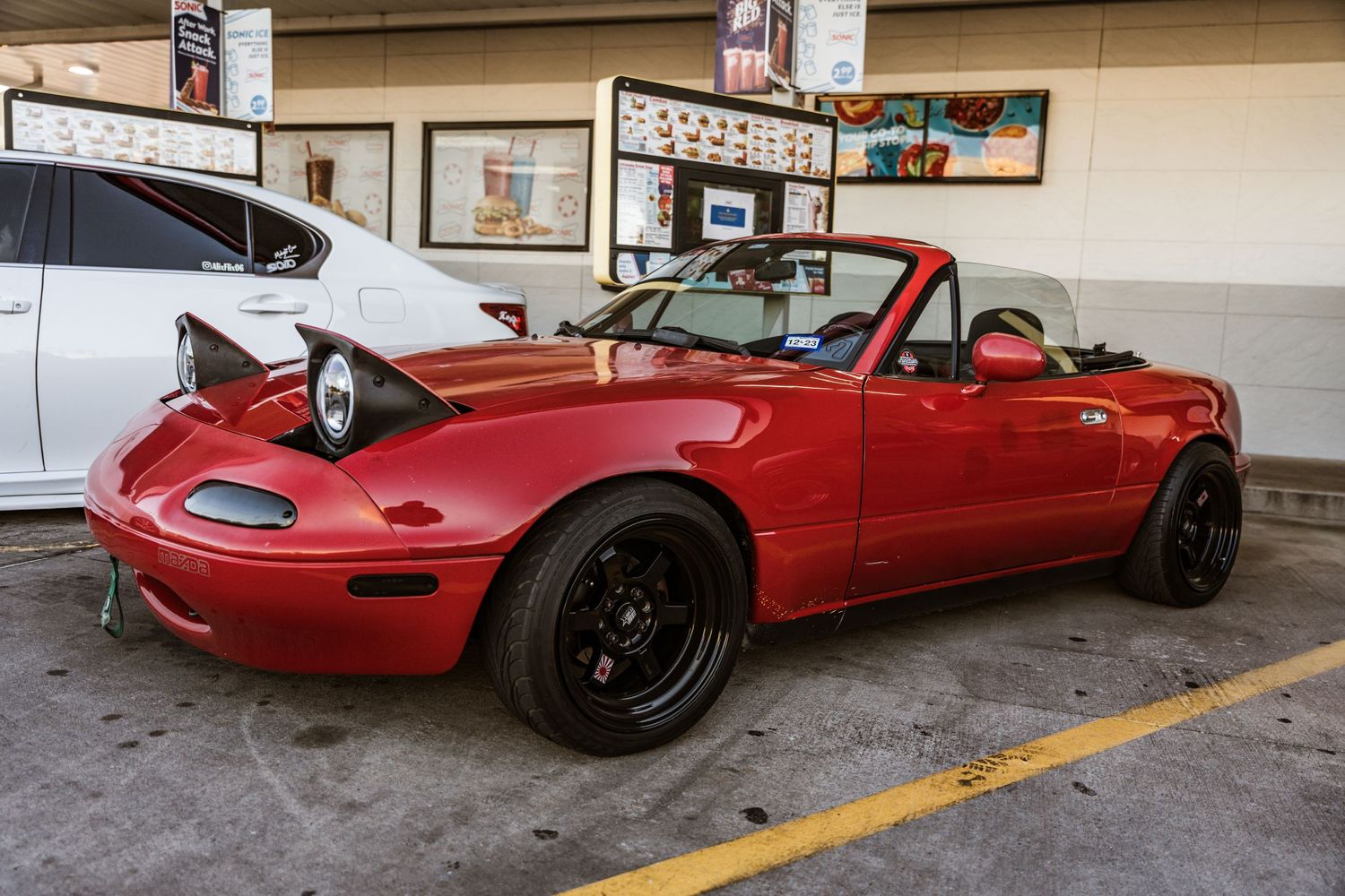 Red Mazda Miata convertible with black wheels parked in front of a store.