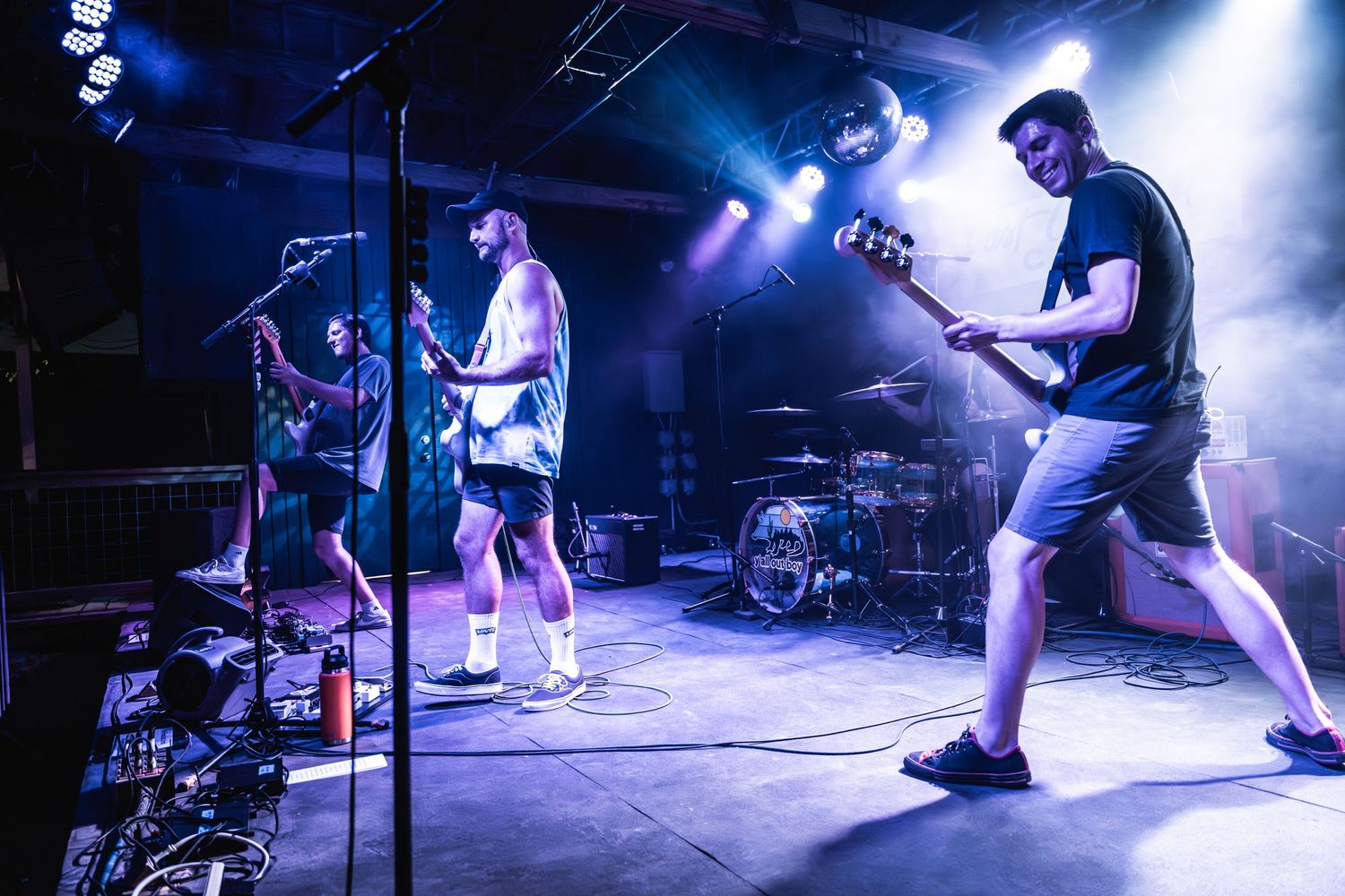 Band members perform on stage with blue lighting and fog effects during an energetic live concert performance.