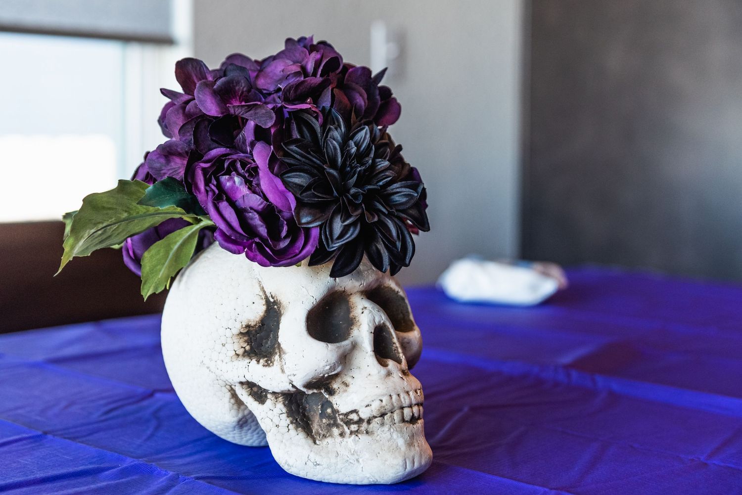 A decorative skull vase filled with deep purple flowers on a blue tablecloth.