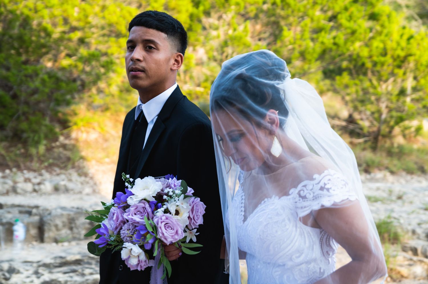 Wedding couple poses outdoors holding purple and white floral bouquet.