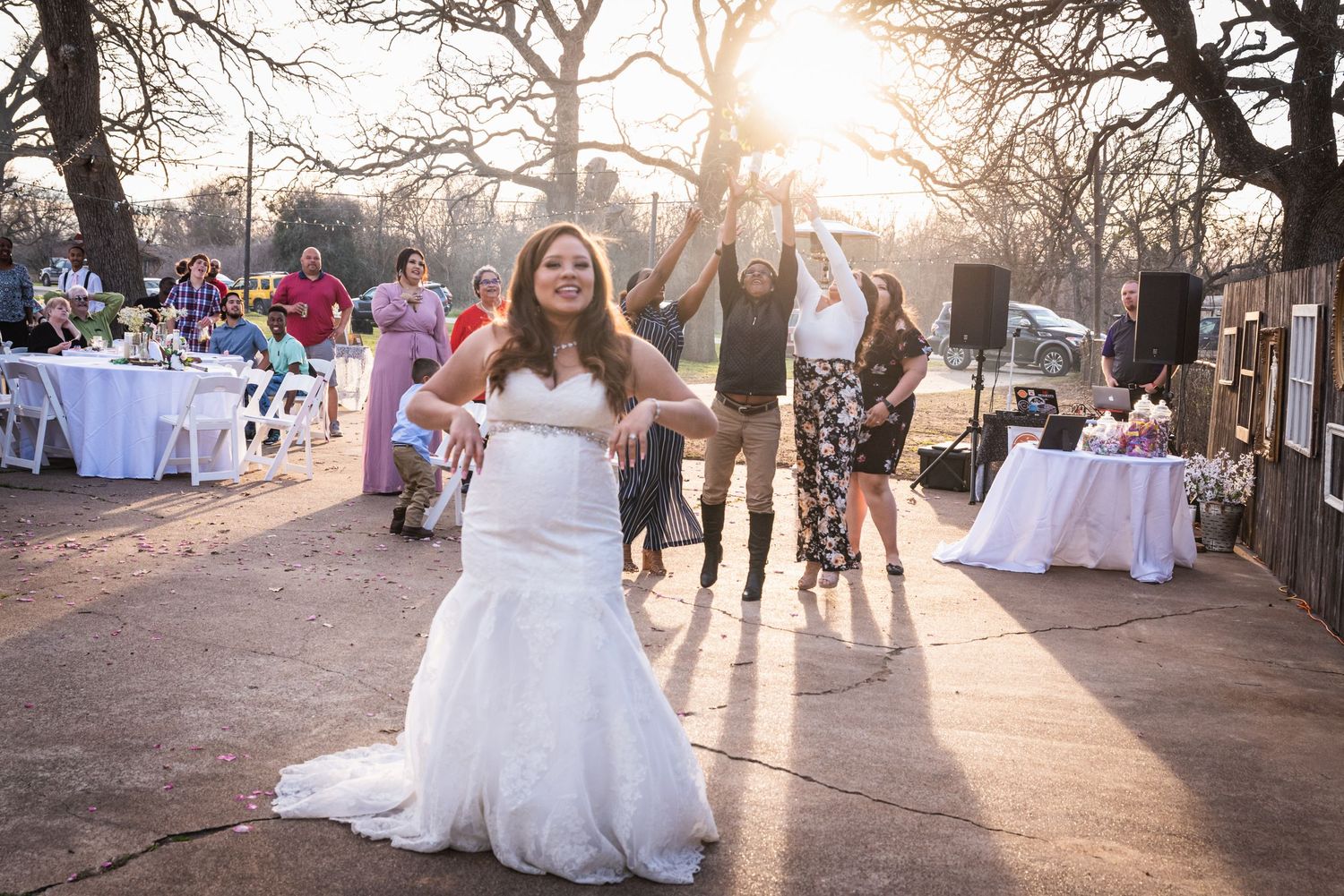 Bride celebrating at sunset with white tables and guests in background at outdoor venue.
