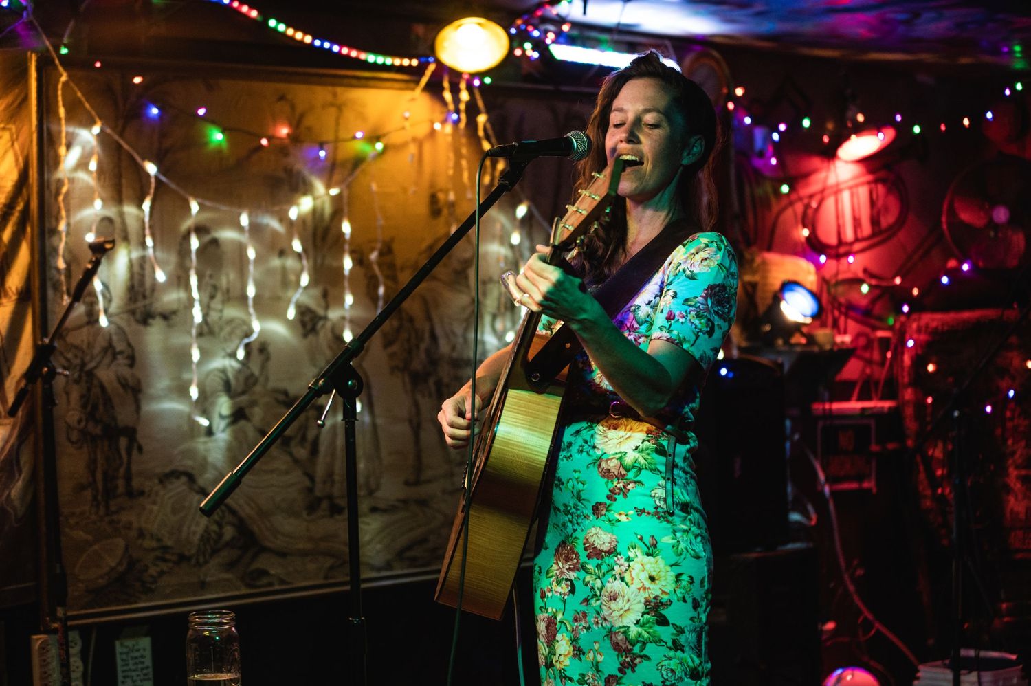 Musician performs with acoustic guitar under colorful stage lights and string light decorations.