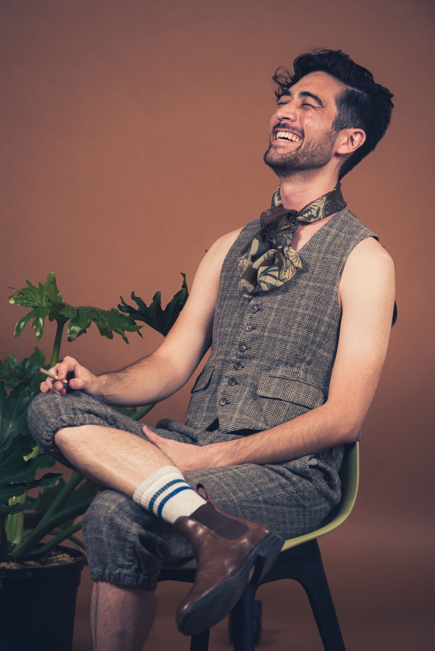 Person in denim overalls sits on chair laughing next to indoor plant against brown backdrop.