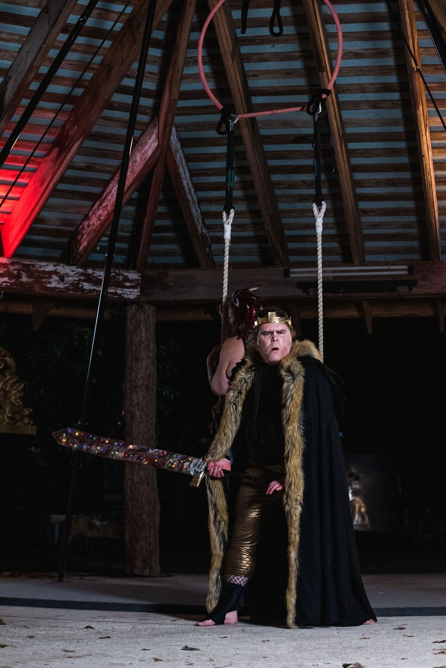 Performer in dramatic cape and costume poses with staff in dimly lit rustic building.