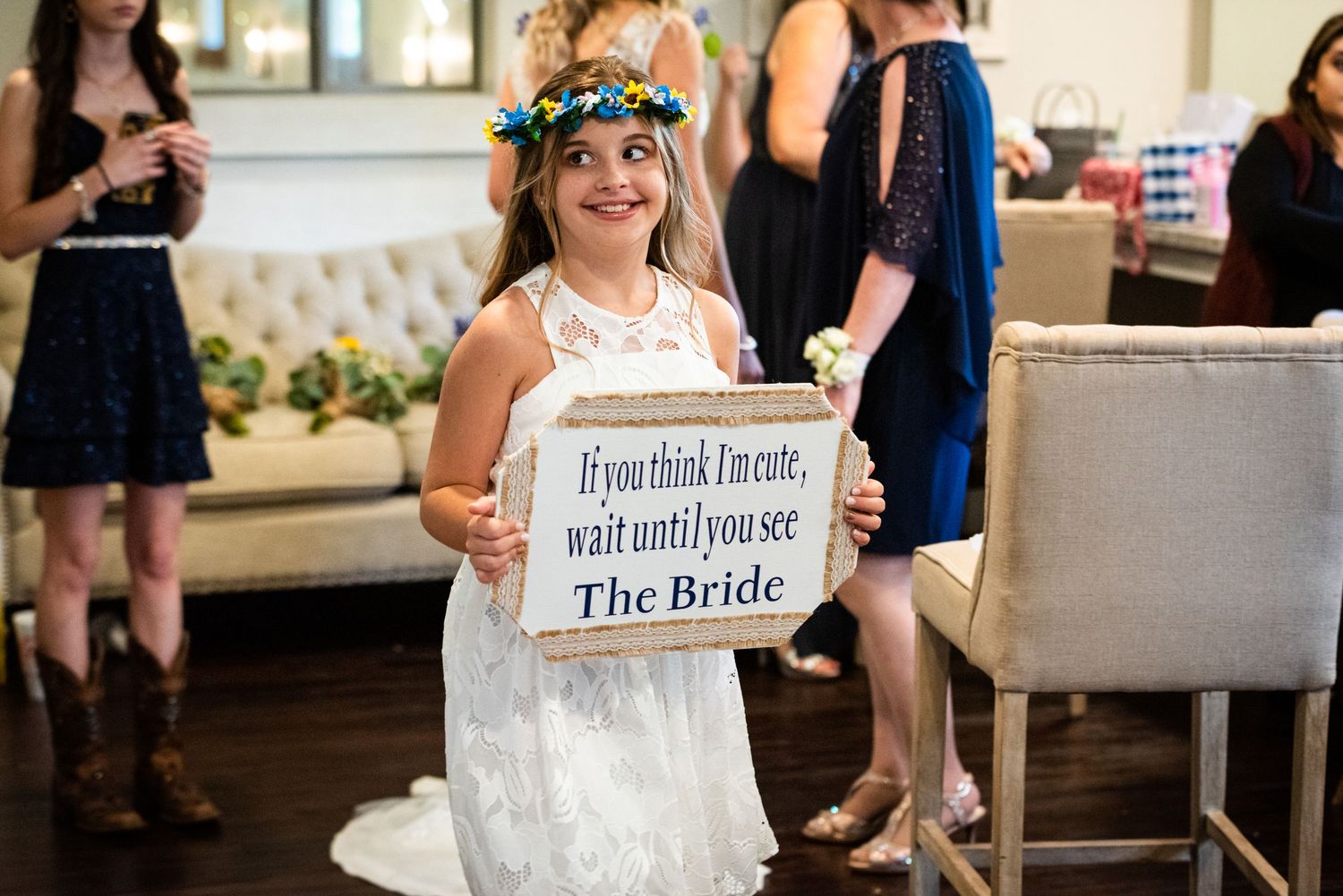 Young flower girl in white dress holds rustic wooden sign while sitting on grey chair.