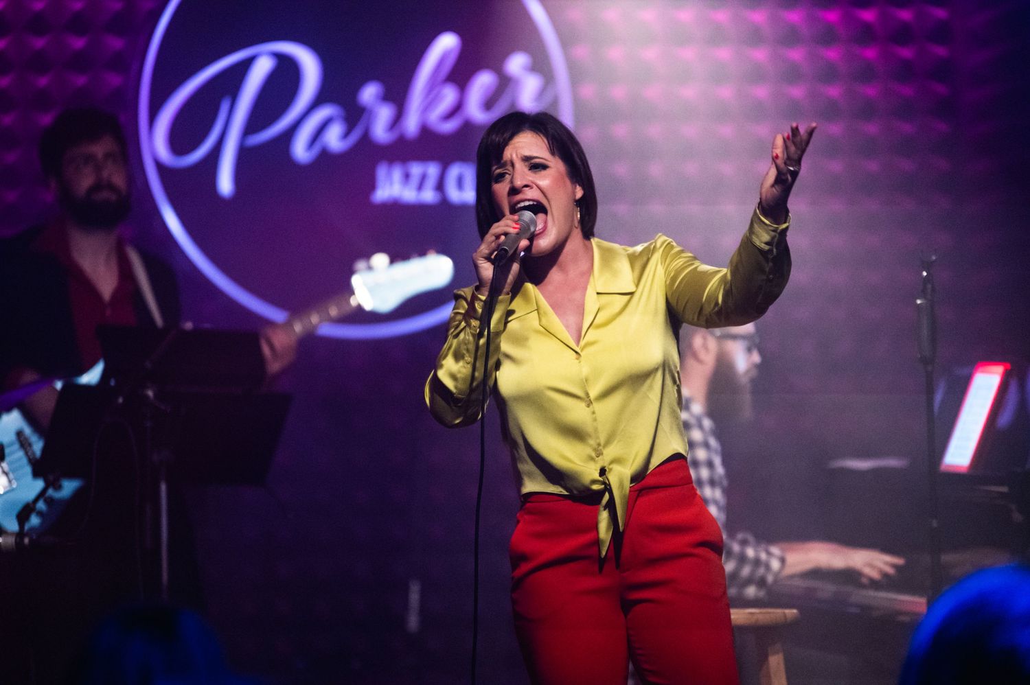 A vocalist performs emotively in yellow attire under the Parker Jazz Club neon sign.