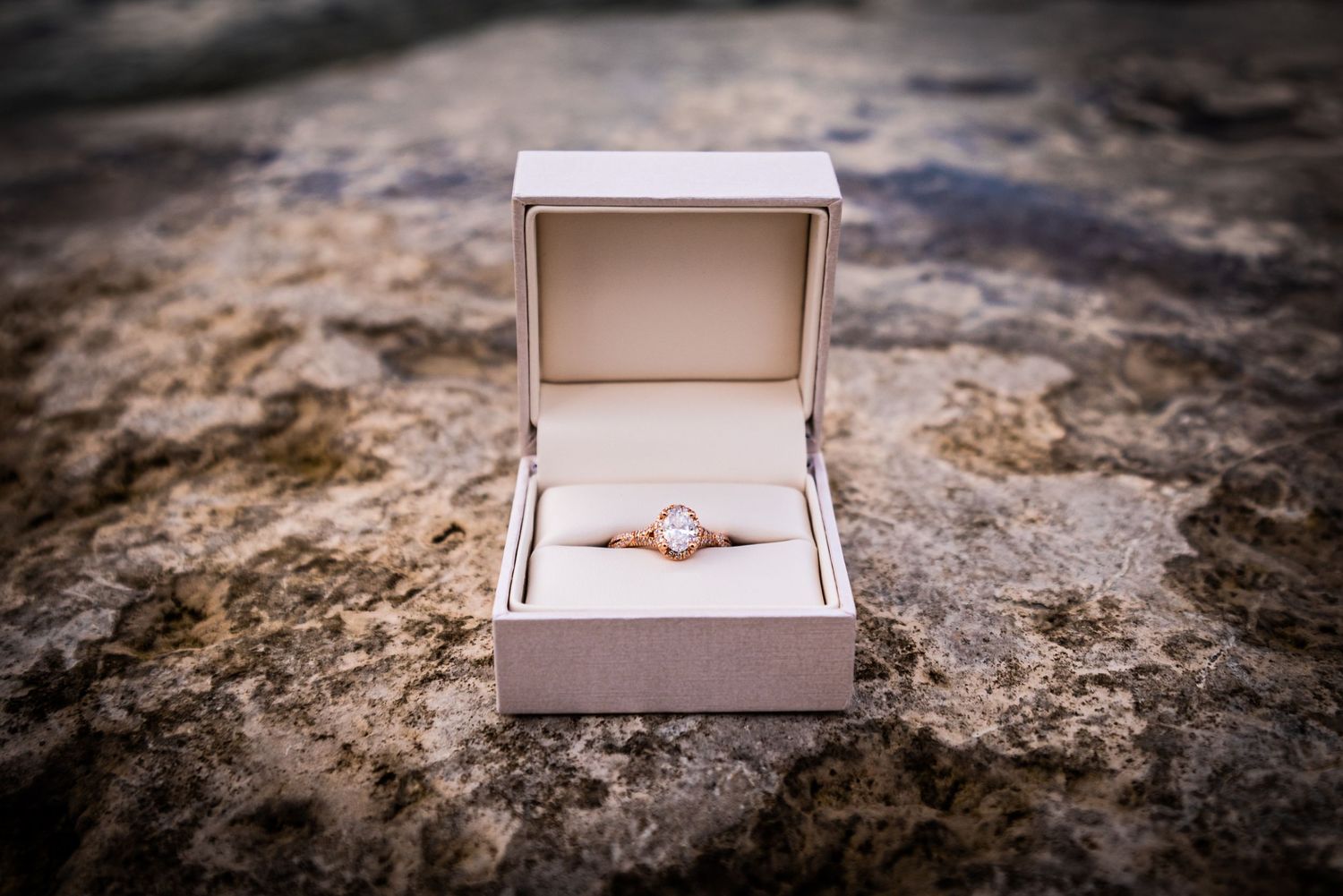 An engagement ring with diamond solitaire sits in an open white ring box on a textured stone surface.