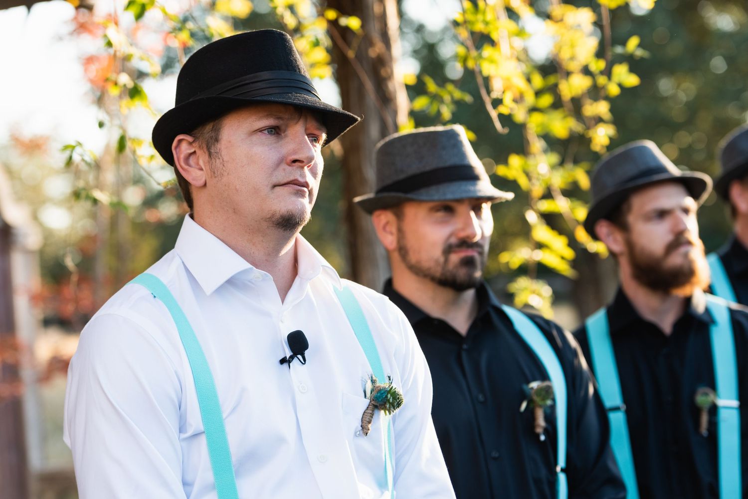 Groomsmen in turquoise suspenders and black hats stand during outdoor ceremony.