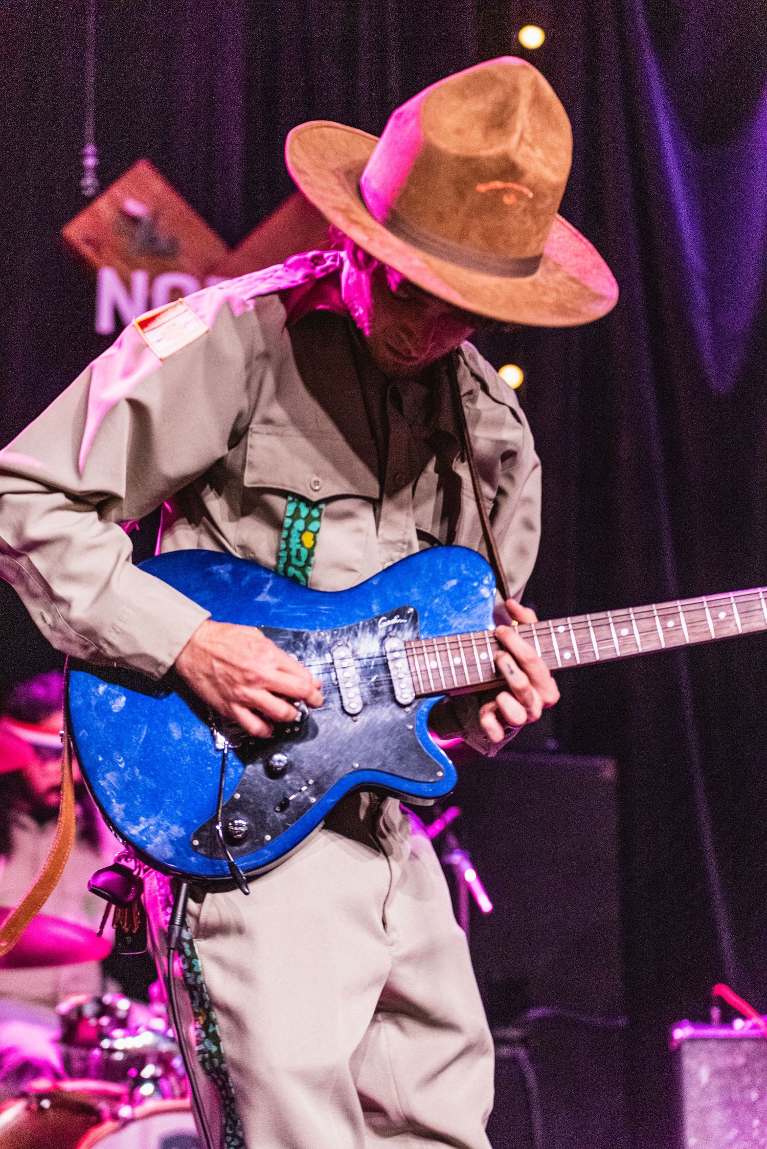 Guitarist in tan western outfit plays blue electric guitar on stage with purple lighting and concert backdrop.