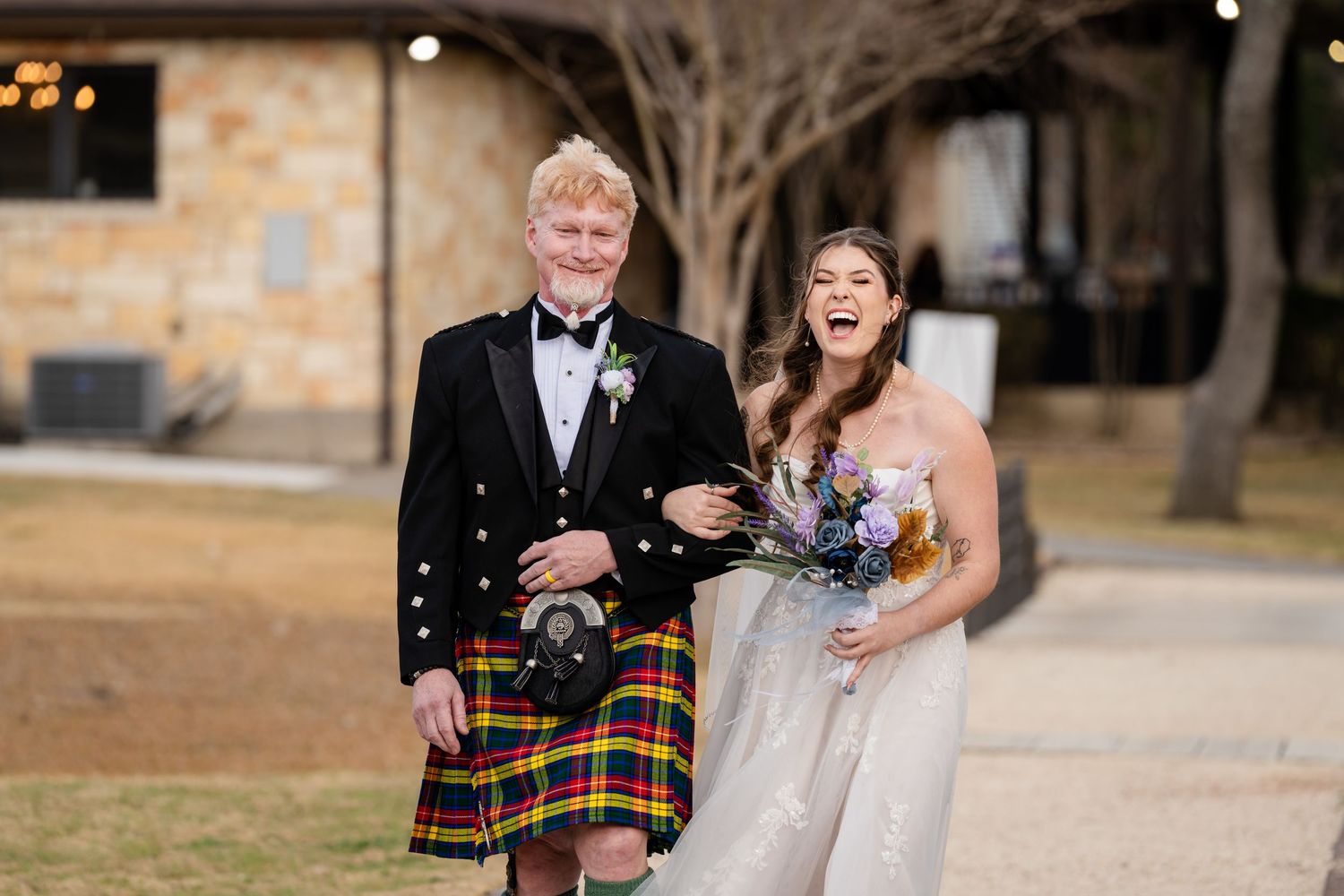 Groom in Scottish tartan kilt and bride in white gown laughing together outdoors at wedding celebration.