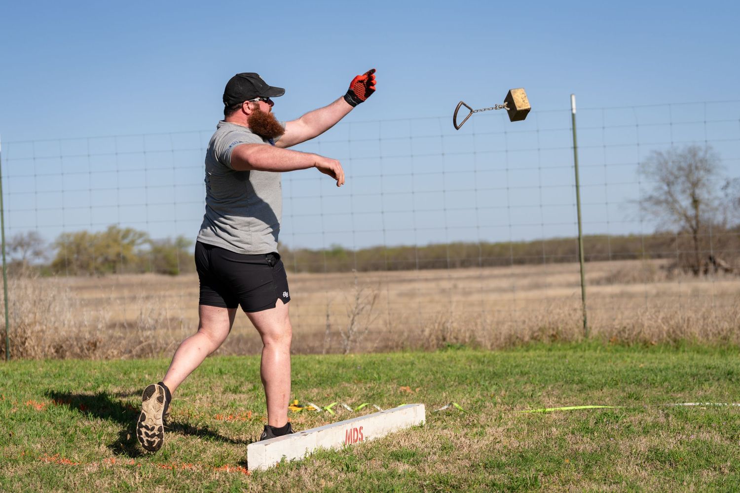 Bearded man in gray tank top throwing a horseshoe on a grass field during an outdoor game on a sunny day.