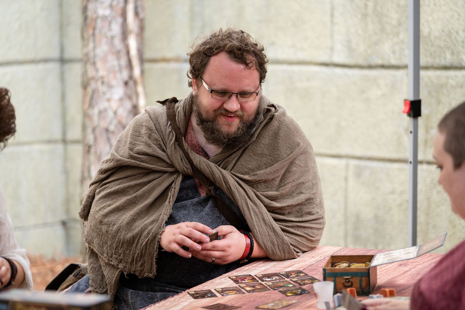 A person in a brown cloak sits at a table while looking at their mobile phone in a medieval-themed setting.