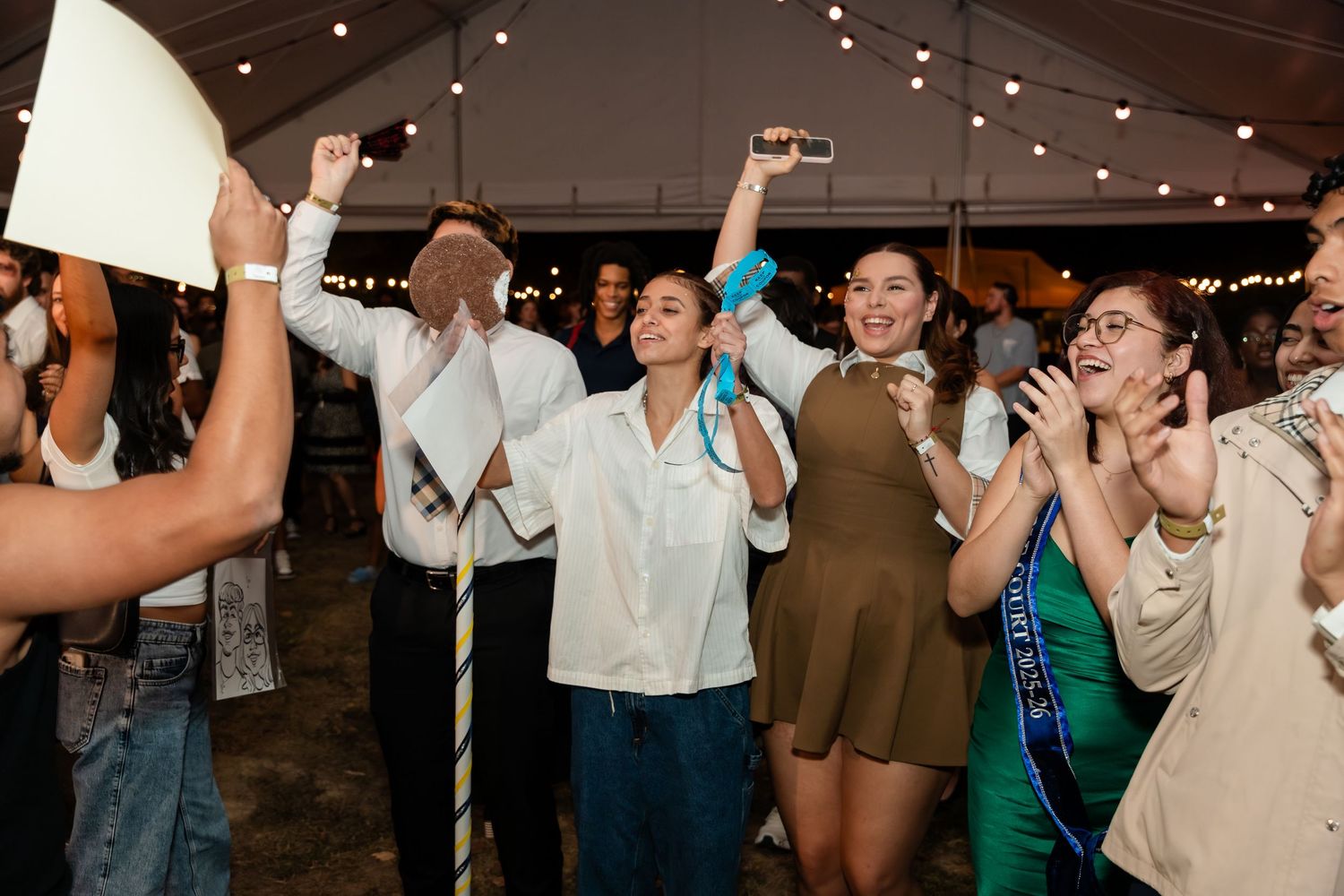 People dancing and celebrating at a lively party with string lights overhead in an indoor venue.