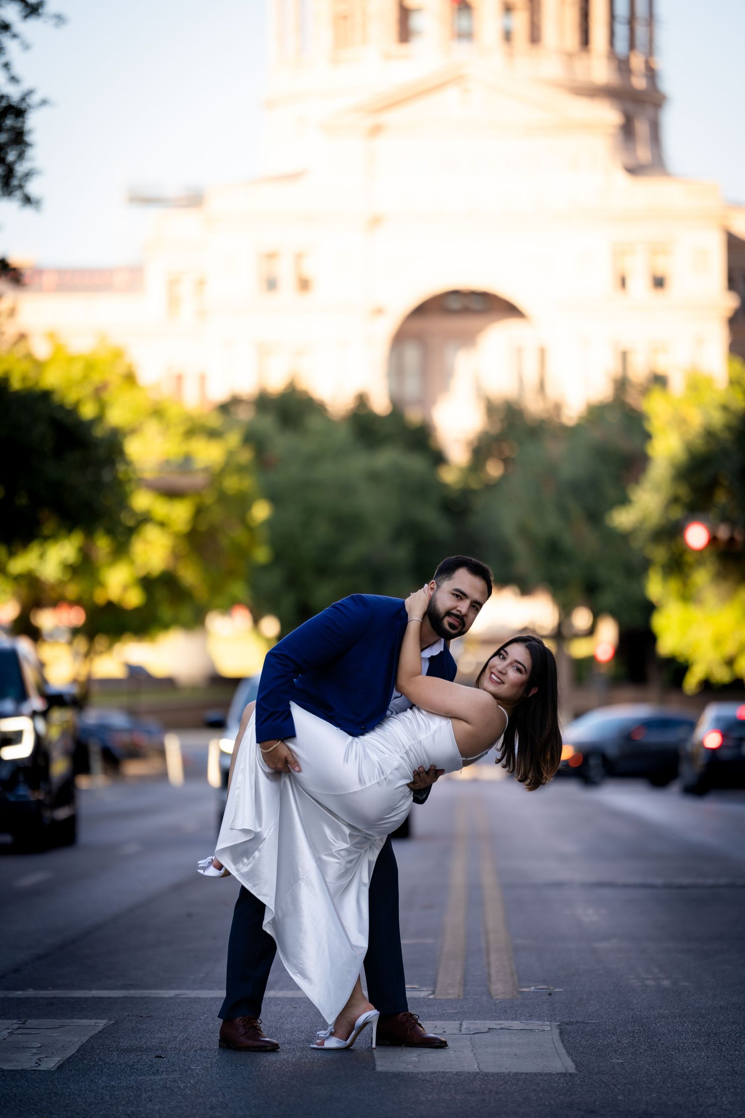 A couple in formal attire dances playfully in an urban street at sunset with buildings in the background.