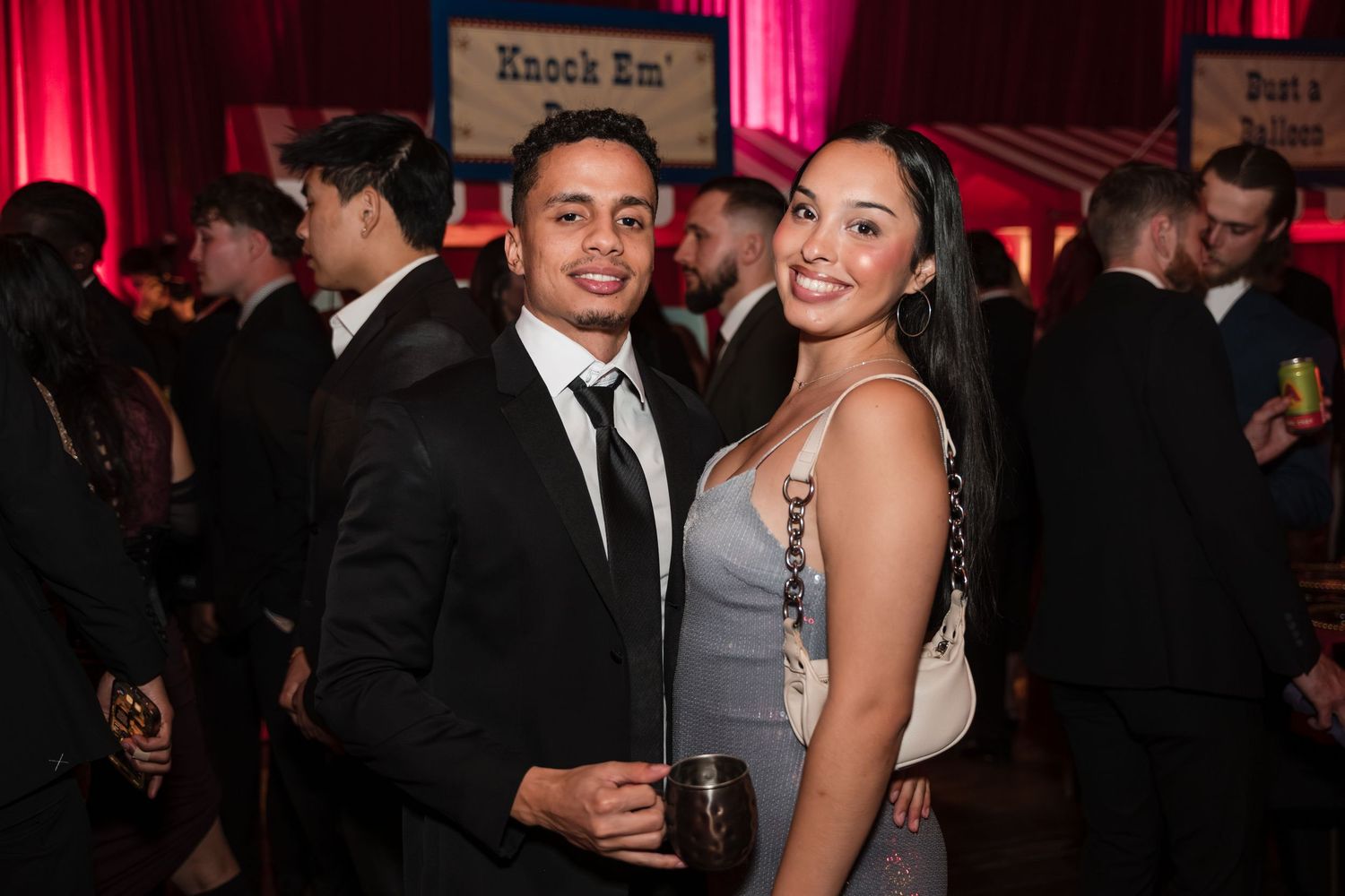 People in formal attire mingle and socialize at an elegant evening event with red lighting and dark ambiance.